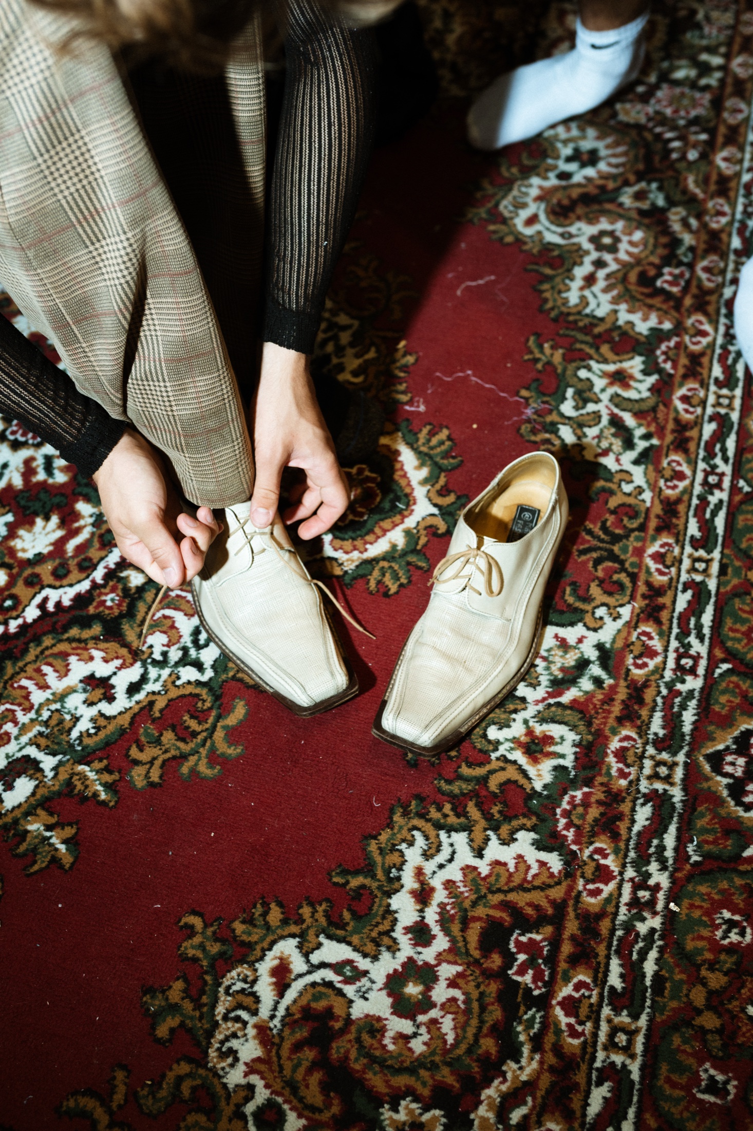 Hands tying a pointed cream shoe on a patterned carpet